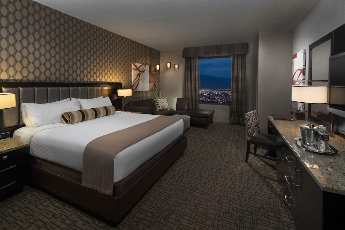 Quarto de hotel com uma cama grande, uma escrivaninha, uma cadeira e vista para a cidade à noite.