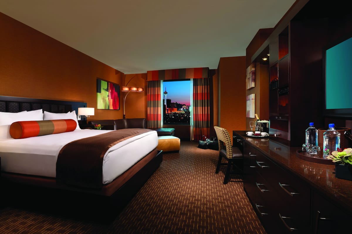 Quarto de hotel com uma cama grande, uma escrivaninha com computador, televisão e vista para a paisagem urbana ao entardecer.