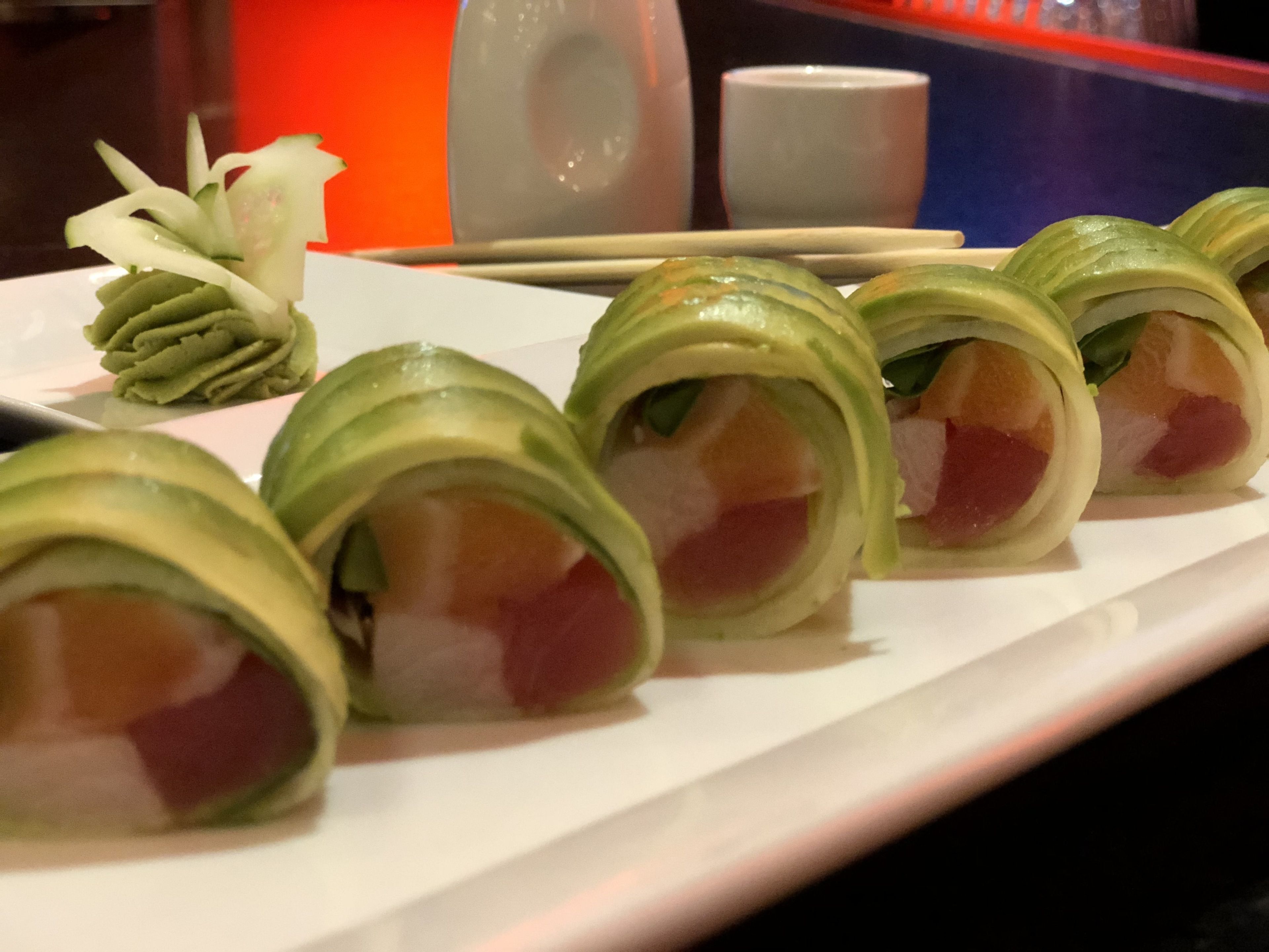 A plate of sushi rolls with a green exterior and a pink interior, accompanied by a small pile of green vegetables and a white cup.