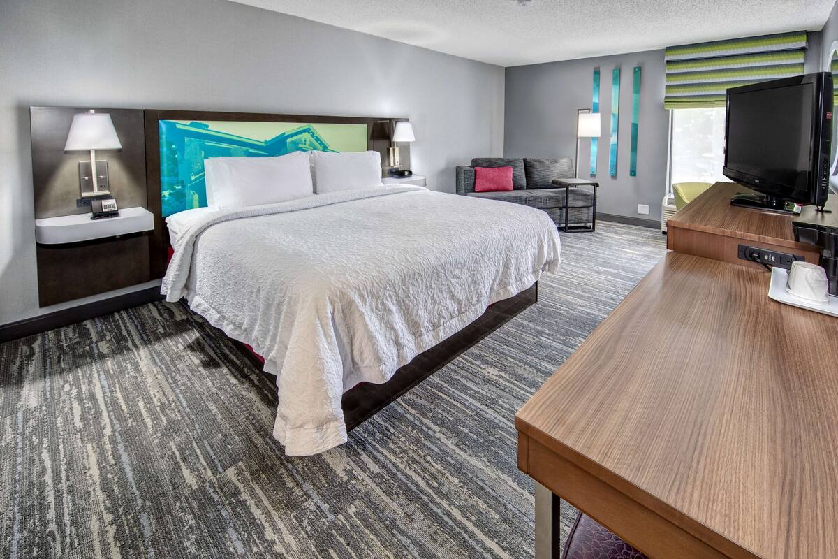 A hotel room with a large bed, a desk, a television, and a window with blinds.