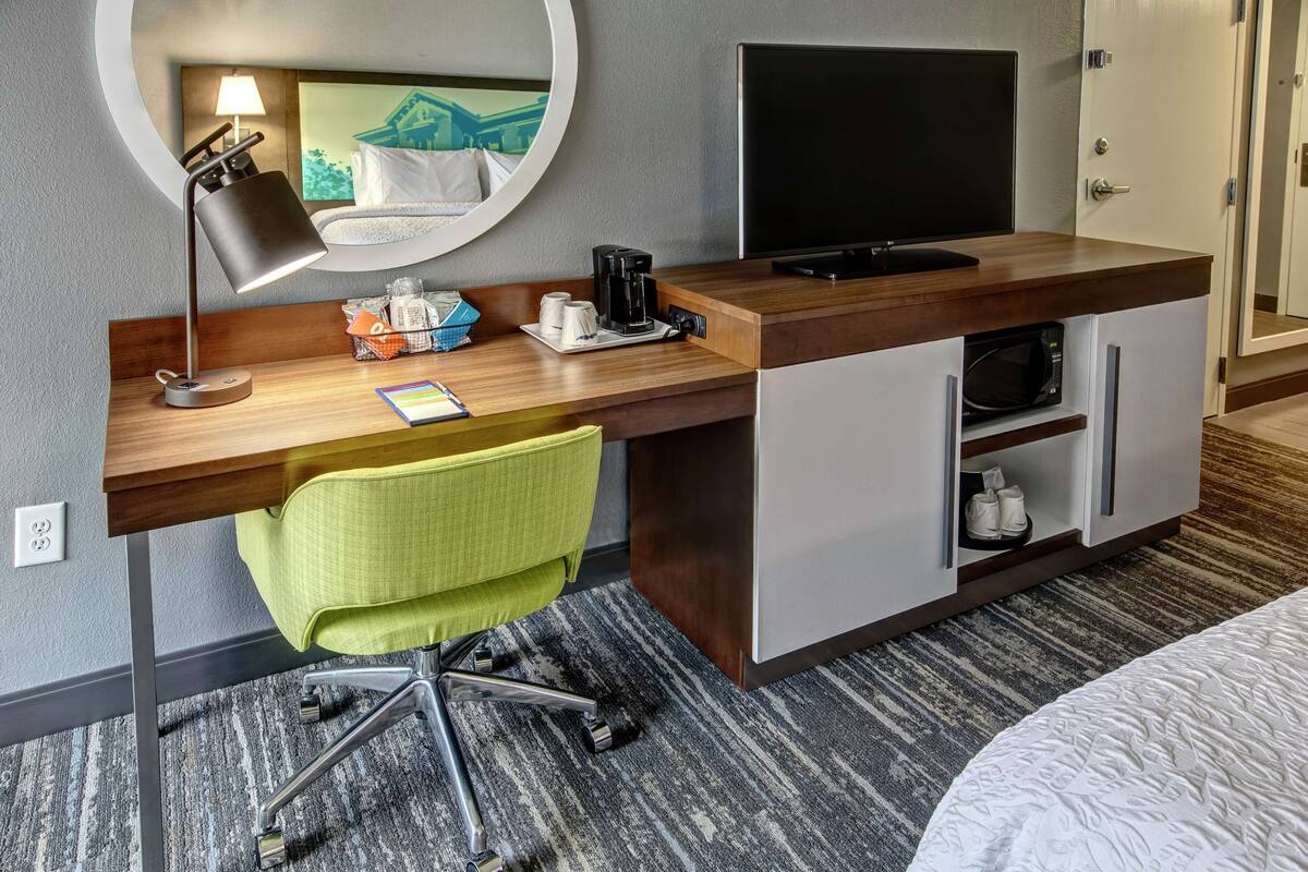 A hotel room with a wooden desk, a green office chair, a flat-screen TV, a coffee maker, and a microwave.