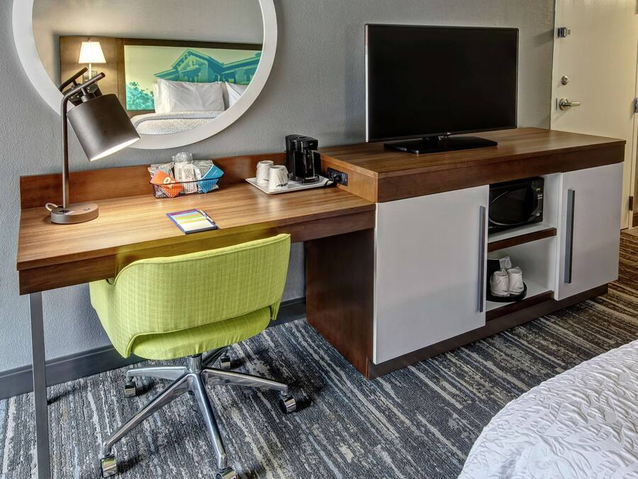 Habitación de hotel con escritorio de madera, una silla de oficina verde, televisor de pantalla plana, cafetera y microondas.