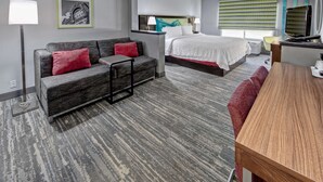 A modern hotel room with a grey sofa, a bed with white bedding, a desk with a computer, and a large window with blinds. - Hampton Inn Batesville (Batesville)