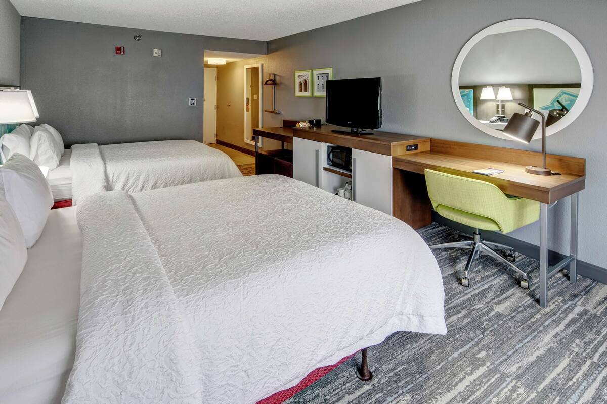 A hotel room with two beds, a desk with a chair, a television, and a round mirror.