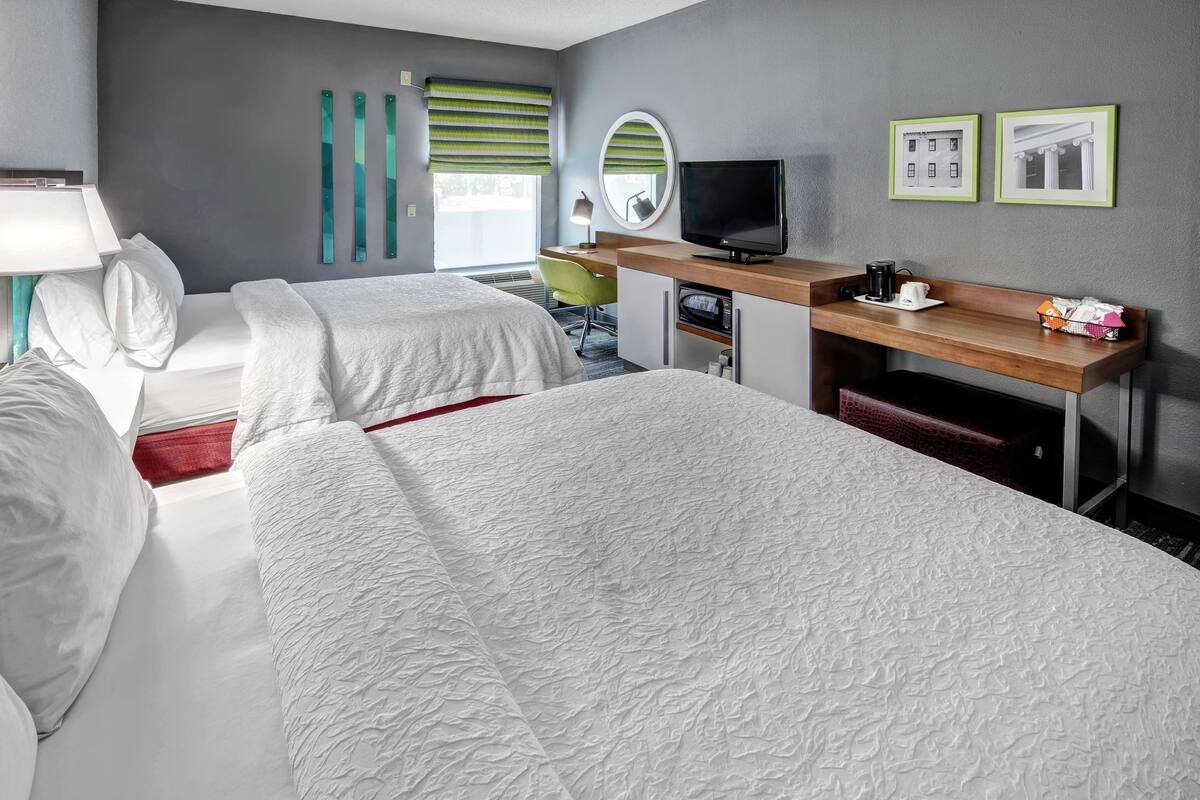 A hotel room with two beds, a desk with a TV, a mirror, and a window with blinds.