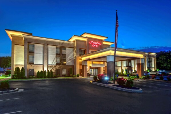 Hampton Inn features a modern architectural design with a flat roof and prominent entrance, illuminated by the hotel's name in neon lights.