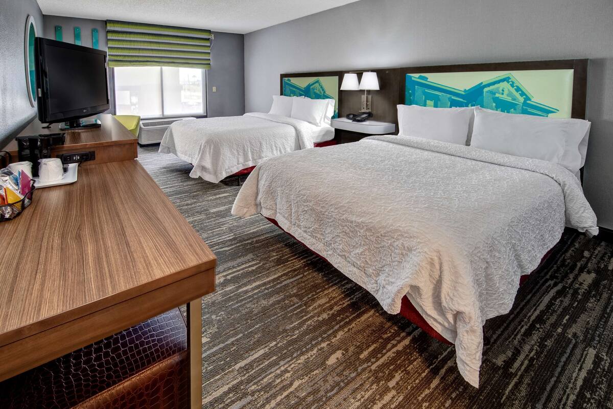 A hotel room with two beds, a desk with a television, a lamp, and artwork on the headboard.