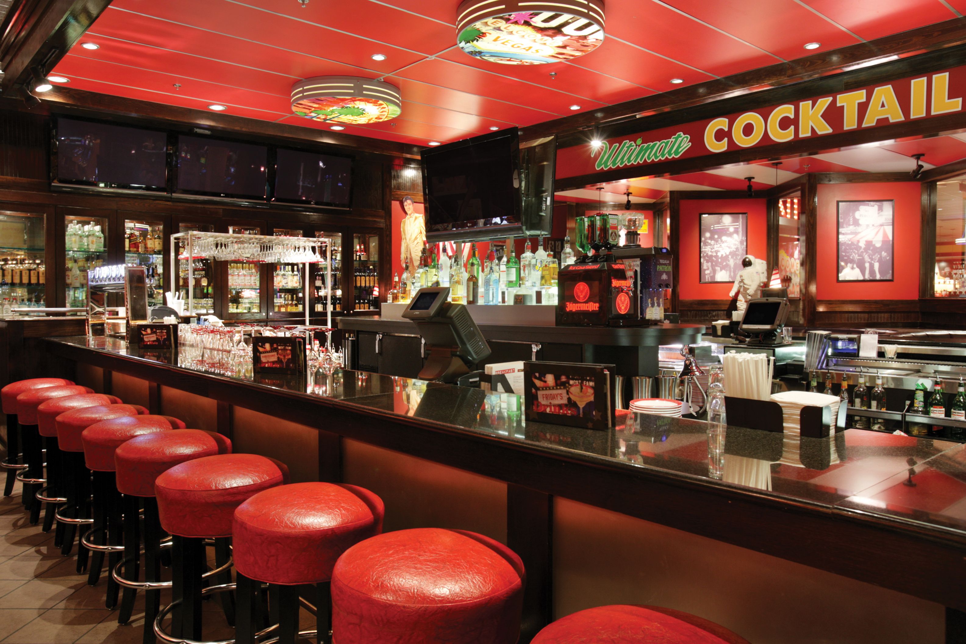Un coin bar avec des tabourets rouges, une enseigne indiquant des cocktails, et une variété de bouteilles et de verres exposés sur des étagères.