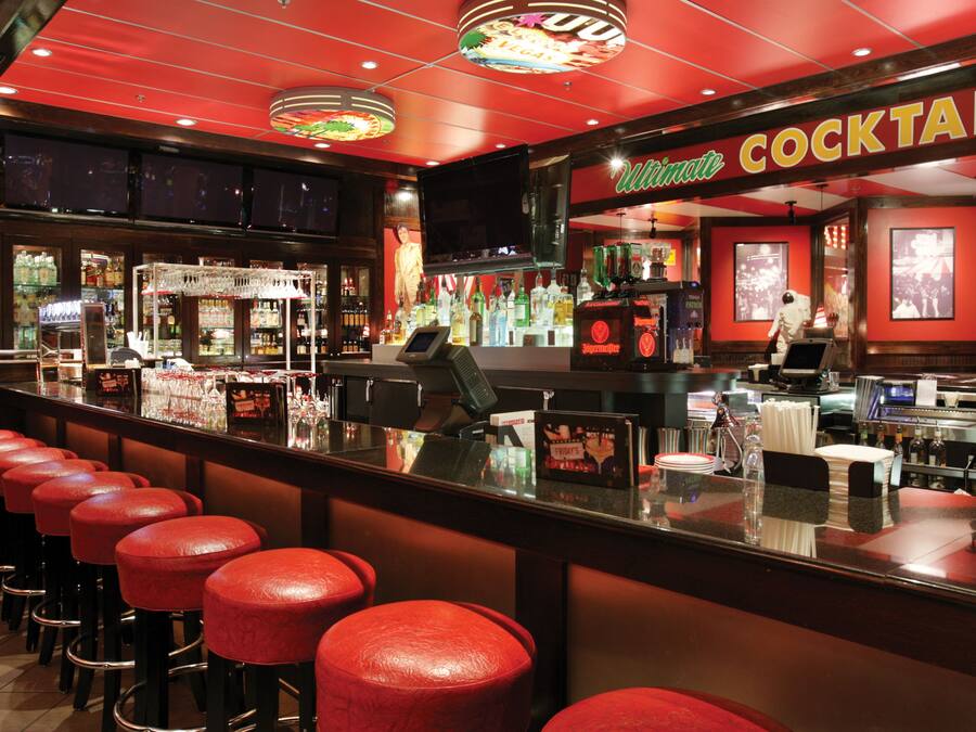 A bar area with red stools, a cocktail sign, and a variety of bottles and glasses on shelves.