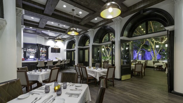 A restaurant with white tablecloths, wooden chairs, and a bar area.
