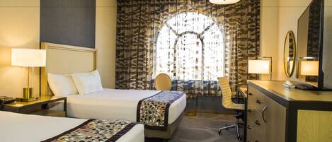 A hotel room with a bed, a desk, a chair, a dresser, and a large window with patterned curtains.