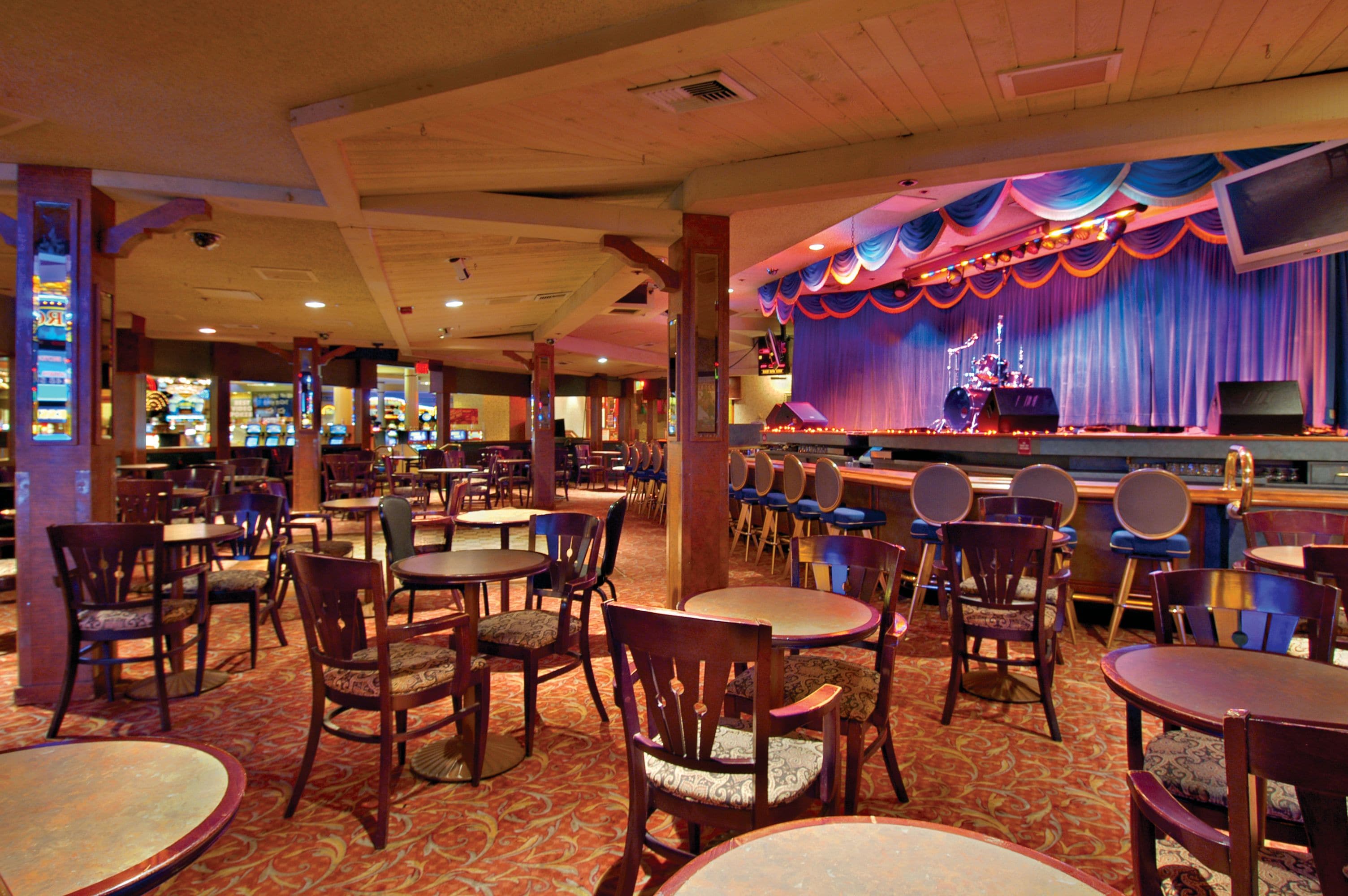 Un restaurante con escenario y zona de bar, que presenta mesas y sillas de madera, además de una alfombra estampada.