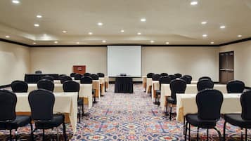 Une salle de conférence avec des rangées de tables et de chaises, un écran de projection et un pupitre.