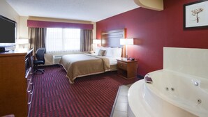 A hotel room with a bed, a desk, a chair, a desk lamp, a bedside table, a nightstand, a TV, a window with curtains, a red carpet, and a white bathtub. - Quality Inn & Suites Stillwater (Stillwater)