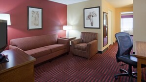 A hotel room with a red sofa, a striped armchair, a desk, and a black office chair. There are two framed pictures on the walls and a lamp on the desk. - Quality Inn & Suites Stillwater (Stillwater)
