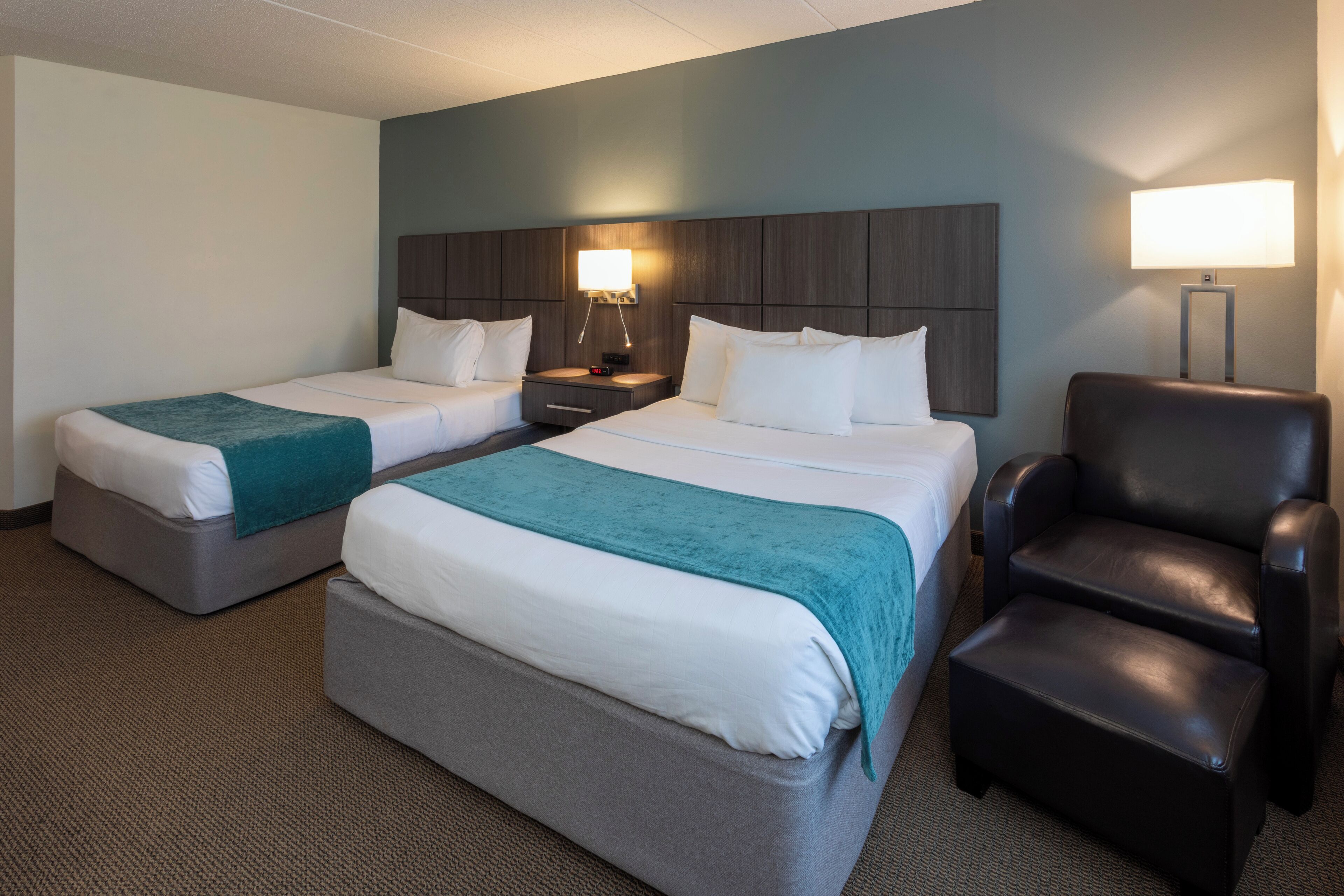 A hotel room with two beds, a desk, a chair, and a lamp.