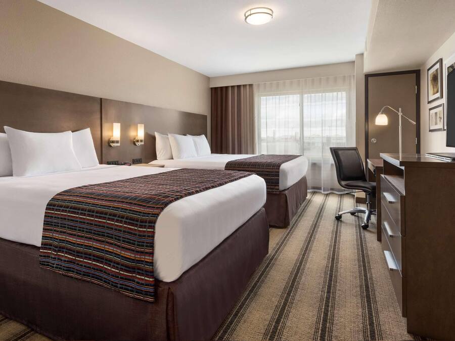A hotel room with two beds, a desk, a chair, a TV, and a large window with curtains.