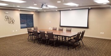 Un salón de conferencias con una gran mesa y sillas, una pantalla de proyector y suelo alfombrado.