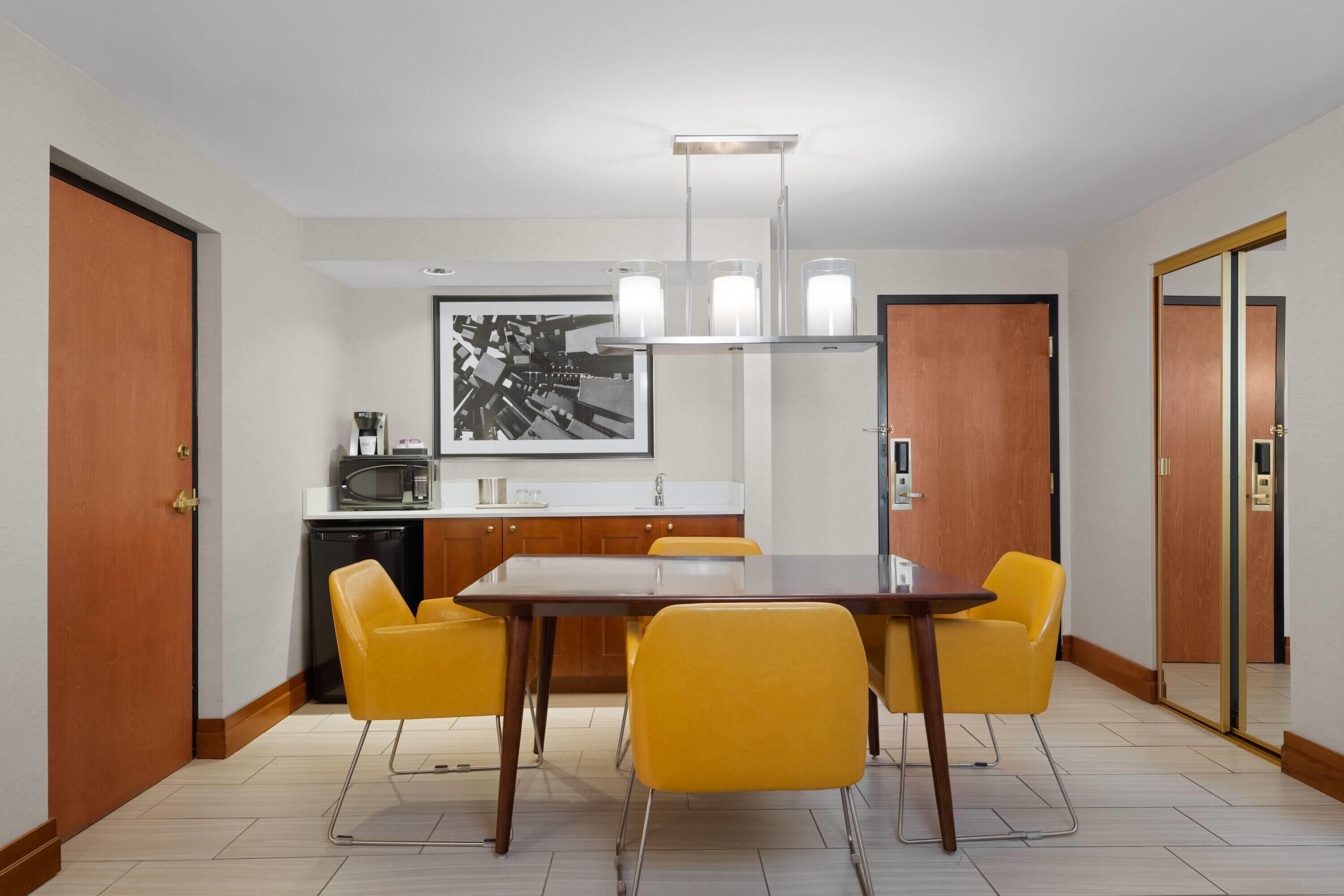 A modern dining area with a wooden table and yellow chairs, a kitchen with a microwave, and a framed cityscape picture on the wall.