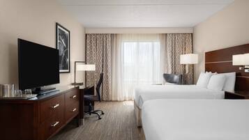 Habitación de hotel con televisión de pantalla plana, escritorio con lámpara, dos camas y un gran ventanal con cortinas.