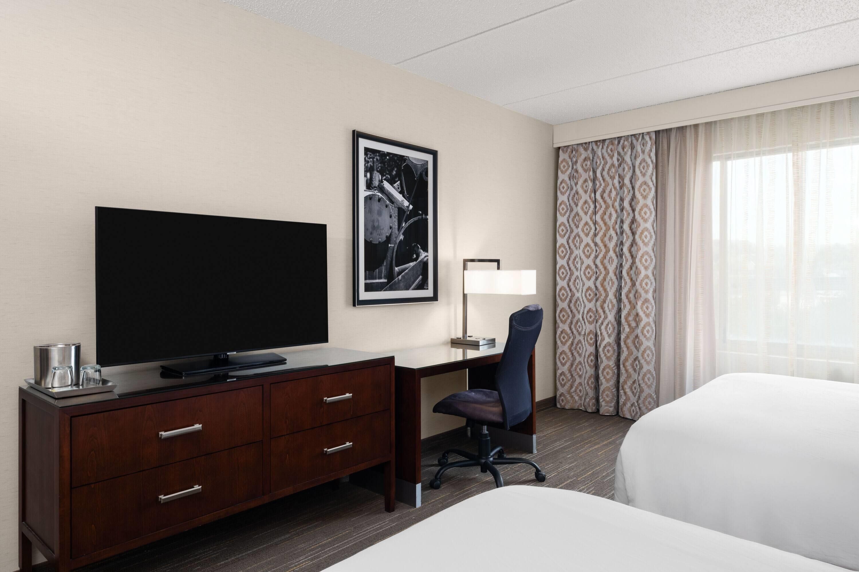 A hotel room with a large flat-screen TV, a desk with a chair, a bed, and a window with patterned curtains.
