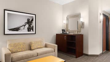 A hotel room with a beige sofa, a yellow ottoman, a wooden cabinet, a framed artwork, and a mirror.