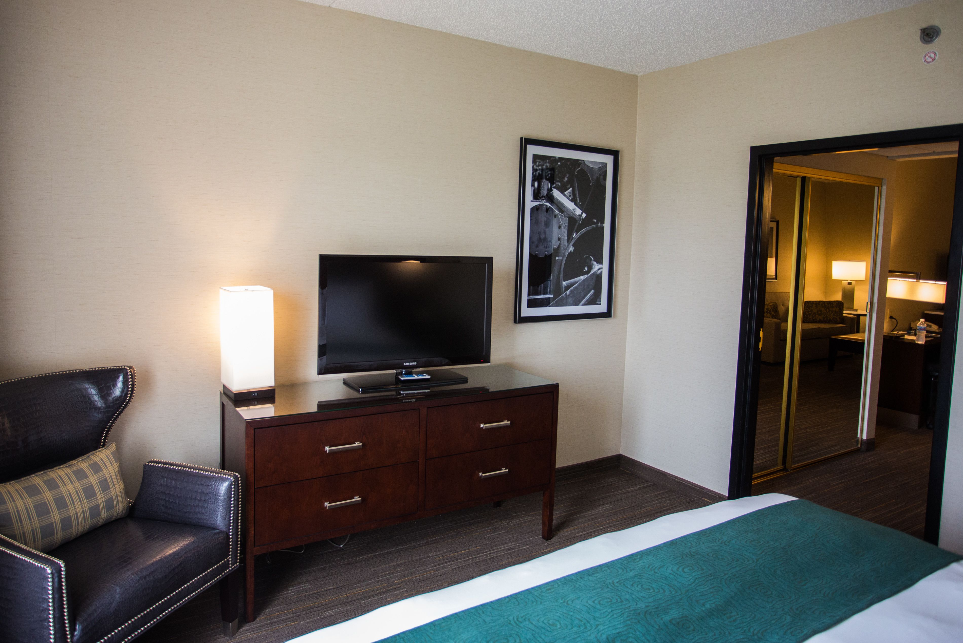 A hotel room with a bed, a TV on a wooden dresser, a chair, and a framed picture on the wall.