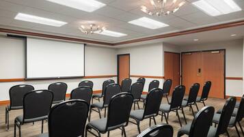 Une salle de conférence avec des rangées de chaises noires, un écran de projection blanc et des panneaux de bois aux murs.