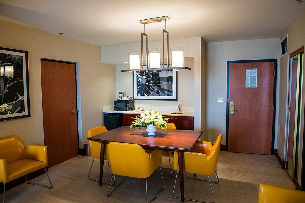 A hotel room with a dining area featuring a wooden table and yellow chairs, a kitchen area with a microwave, and a framed artwork on the wall.
