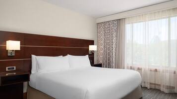 A hotel room with a large bed, two bedside lamps, a nightstand, and a window with sheer curtains.