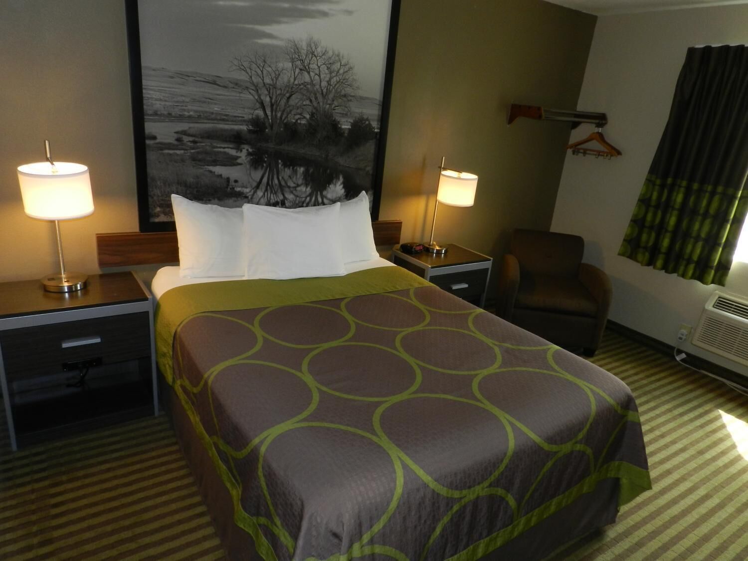 A hotel room with a large bed, two bedside tables with lamps, a chair, a coat rack, and a large framed picture on the wall.