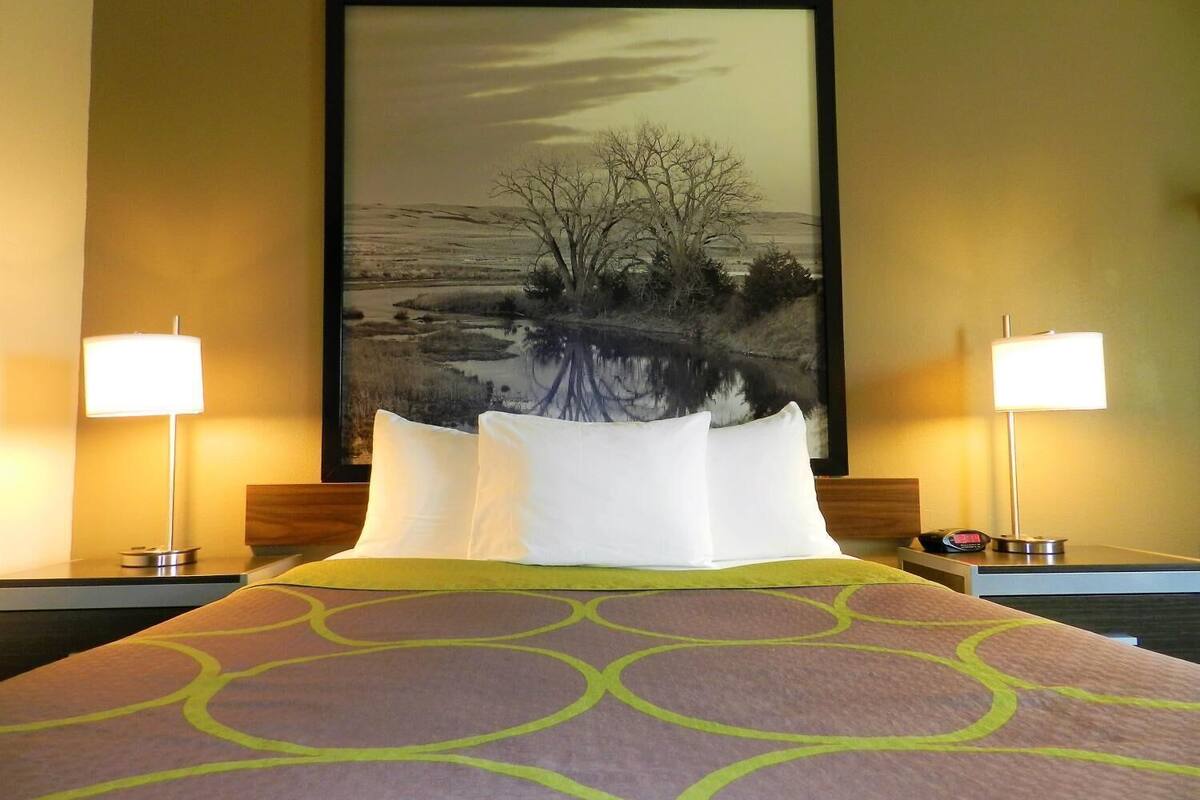 A hotel room with a large bed, two bedside lamps, a framed landscape painting, and a patterned bedspread.
