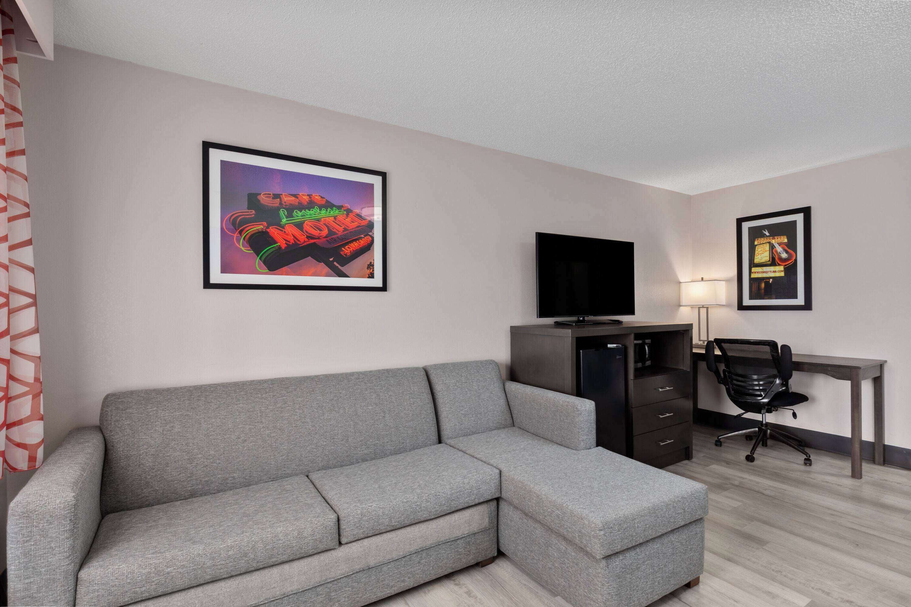 A modern room with a grey sectional sofa, a flat-screen TV on a cabinet, a desk with a chair, and two framed posters on the wall.