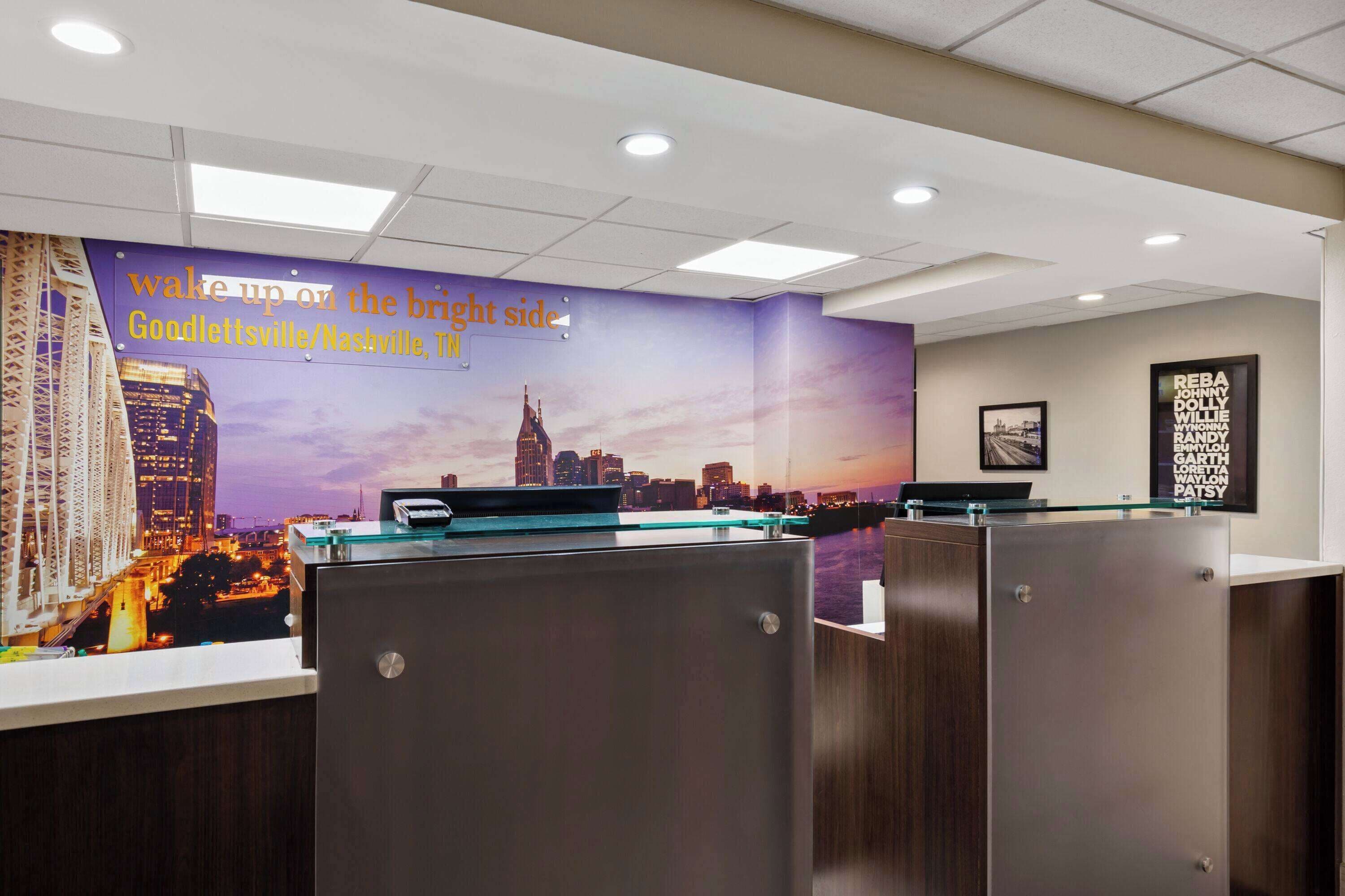 A hotel reception area with a cityscape mural, a reception desk, and a framed sign listing names.