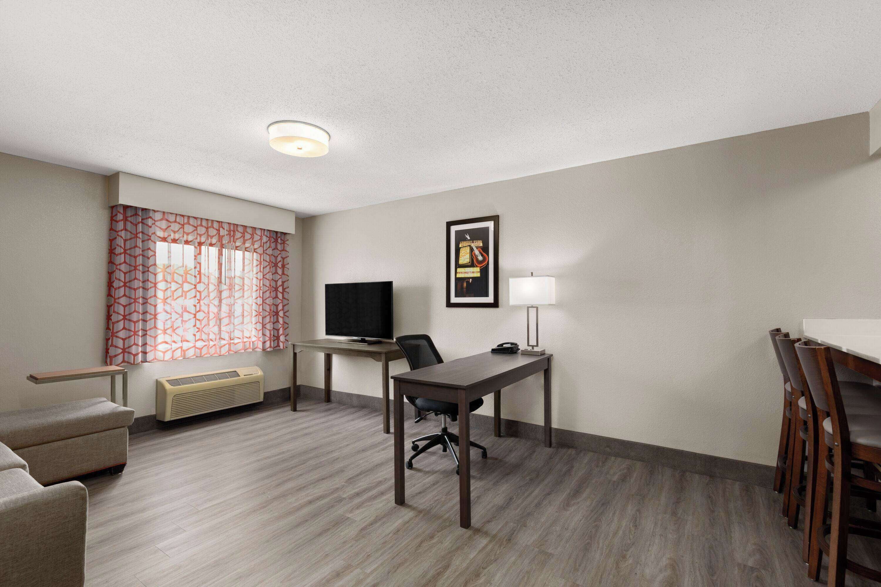 A hotel room with a desk, chair, TV, and a small table with a lamp.