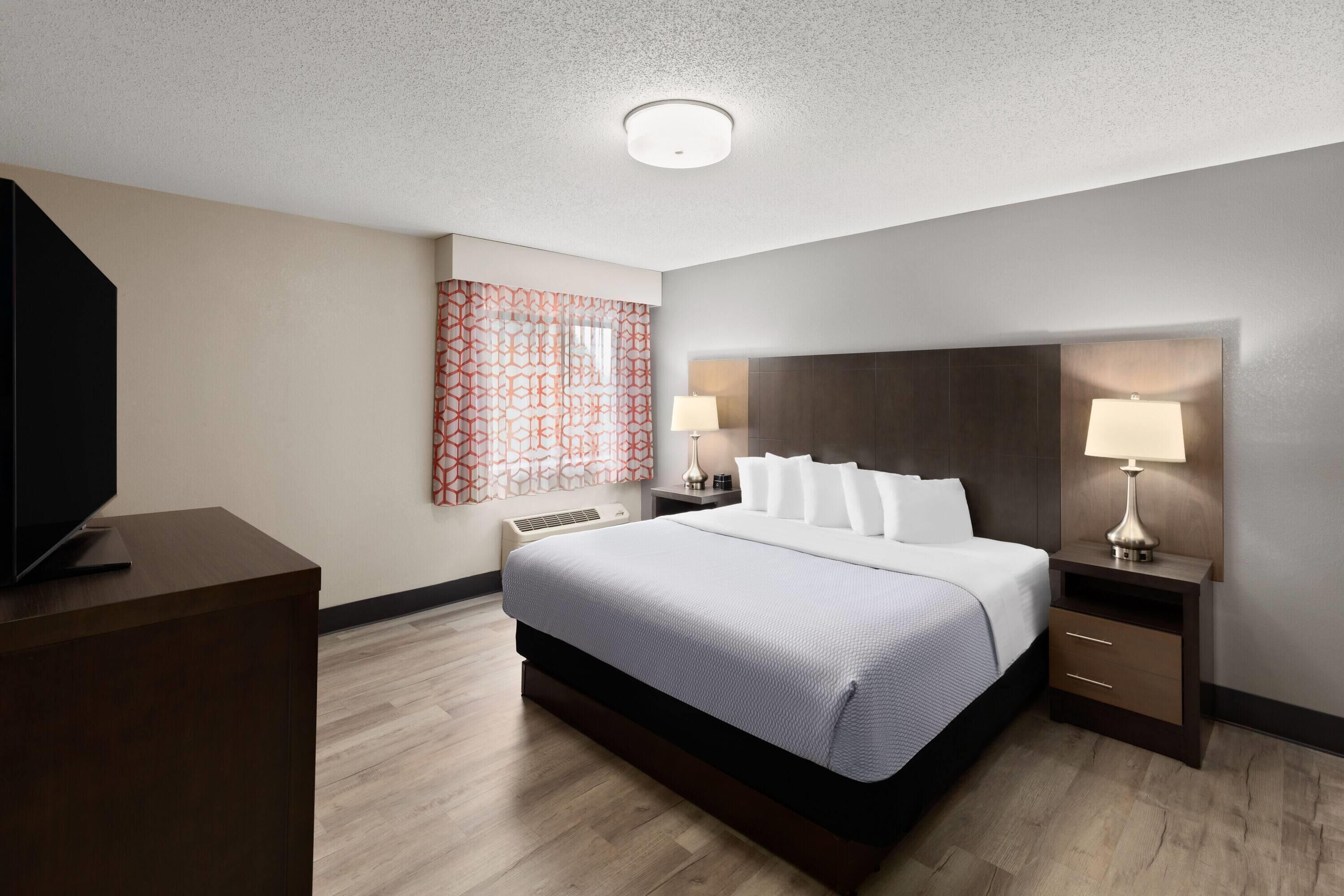 A modern hotel room with a large bed, bedside tables with lamps, a flat-screen TV, and a patterned window treatment.