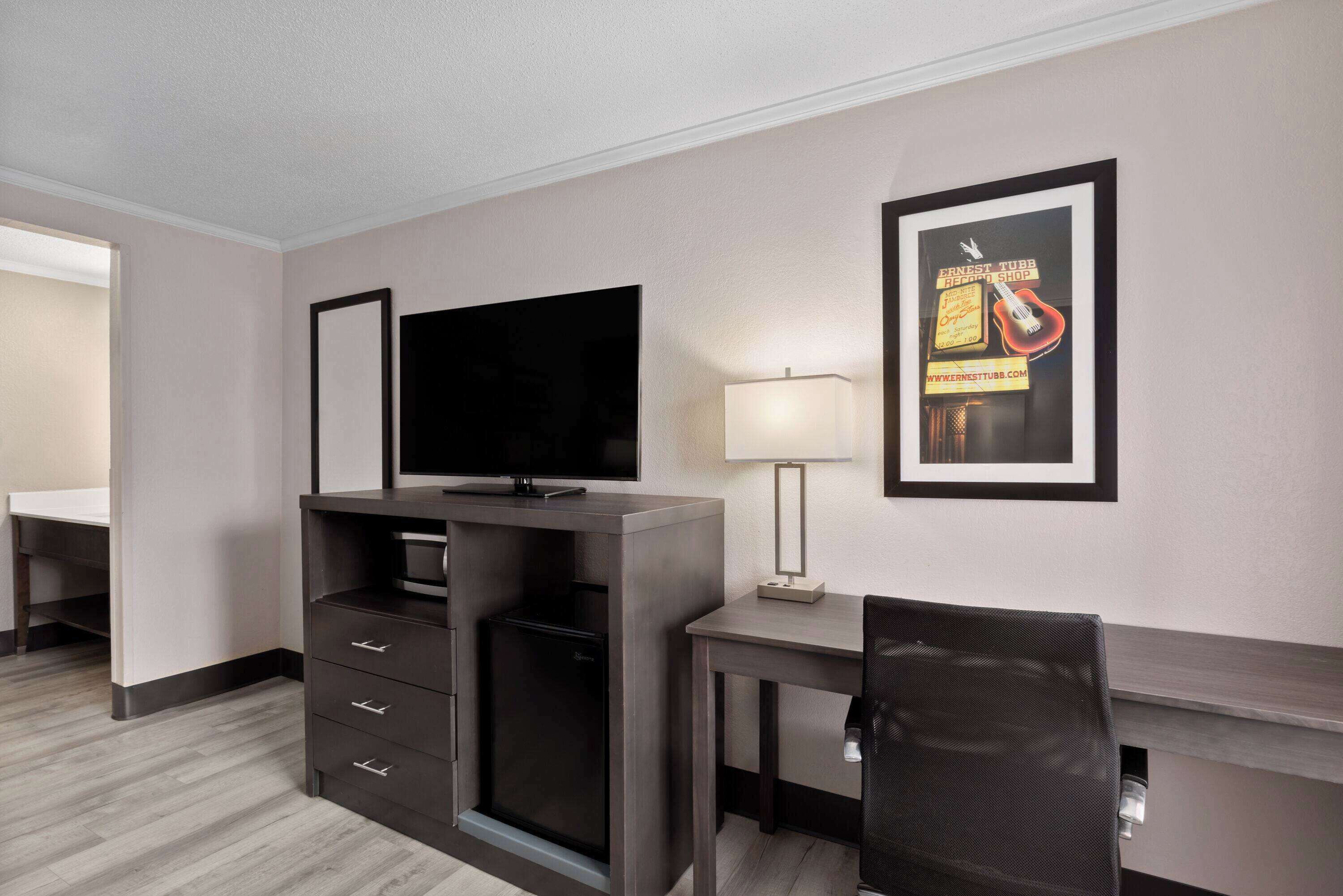 Una camera d'albergo con televisione a schermo piatto, una scrivania con lampada, una sedia e un quadro appeso al muro.