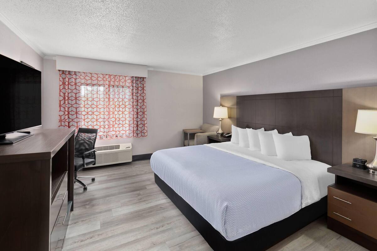 Ein Hotelzimmer mit einem großen Bett, einem Schreibtisch, einem Sessel, einem Fernseher und einem Fenster mit gemusterten Vorhängen.