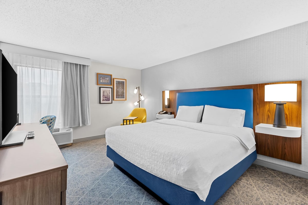 A hotel room with a large bed, a desk, a chair, and a nightstand with a lamp.