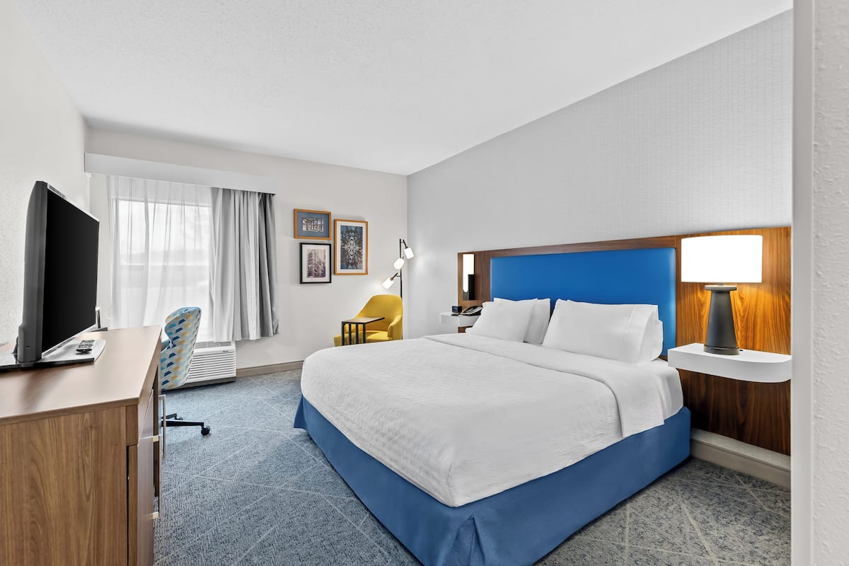 A hotel room with a bed, a desk with a computer, a chair, a lamp, and a window with curtains.