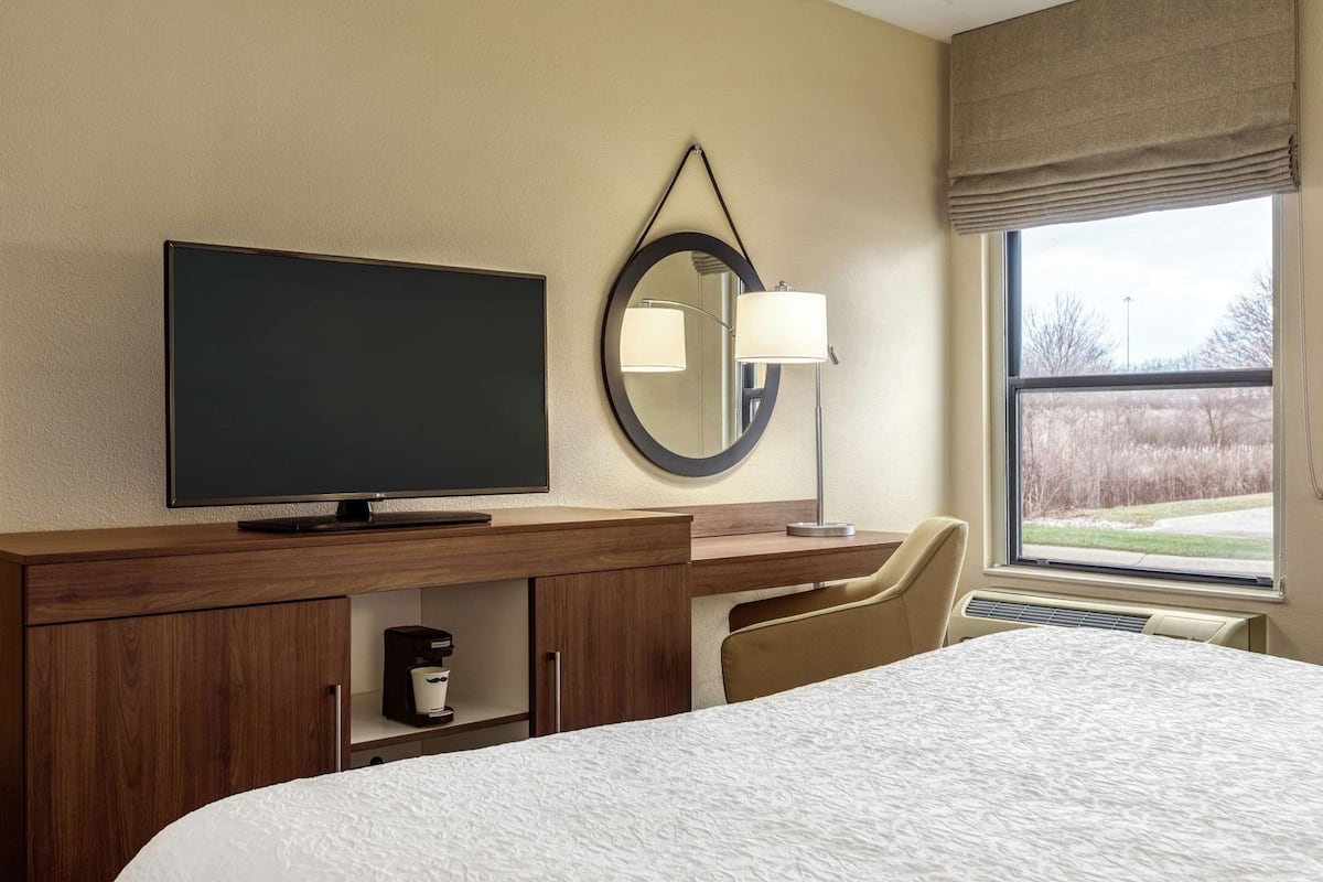 A hotel room with a bed, a TV, a desk with a lamp, a chair, and a window with a view.