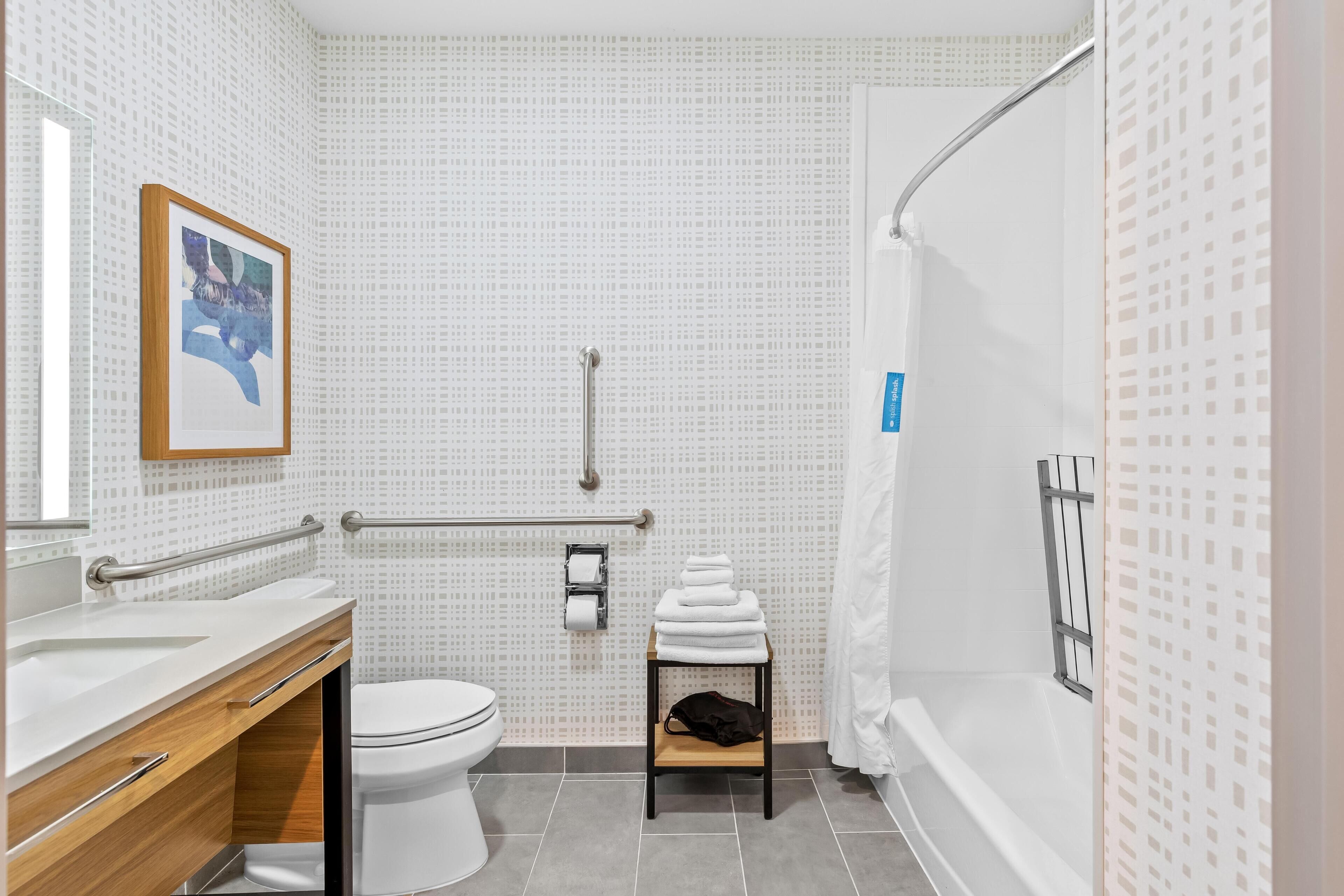 A modern bathroom with a white sink, toilet, and shower area. There is a towel rack with folded towels and a small table with a black bag.