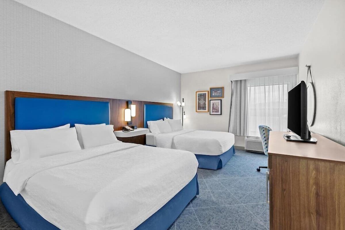 A hotel room with two beds, a desk, a TV, and a window with curtains.