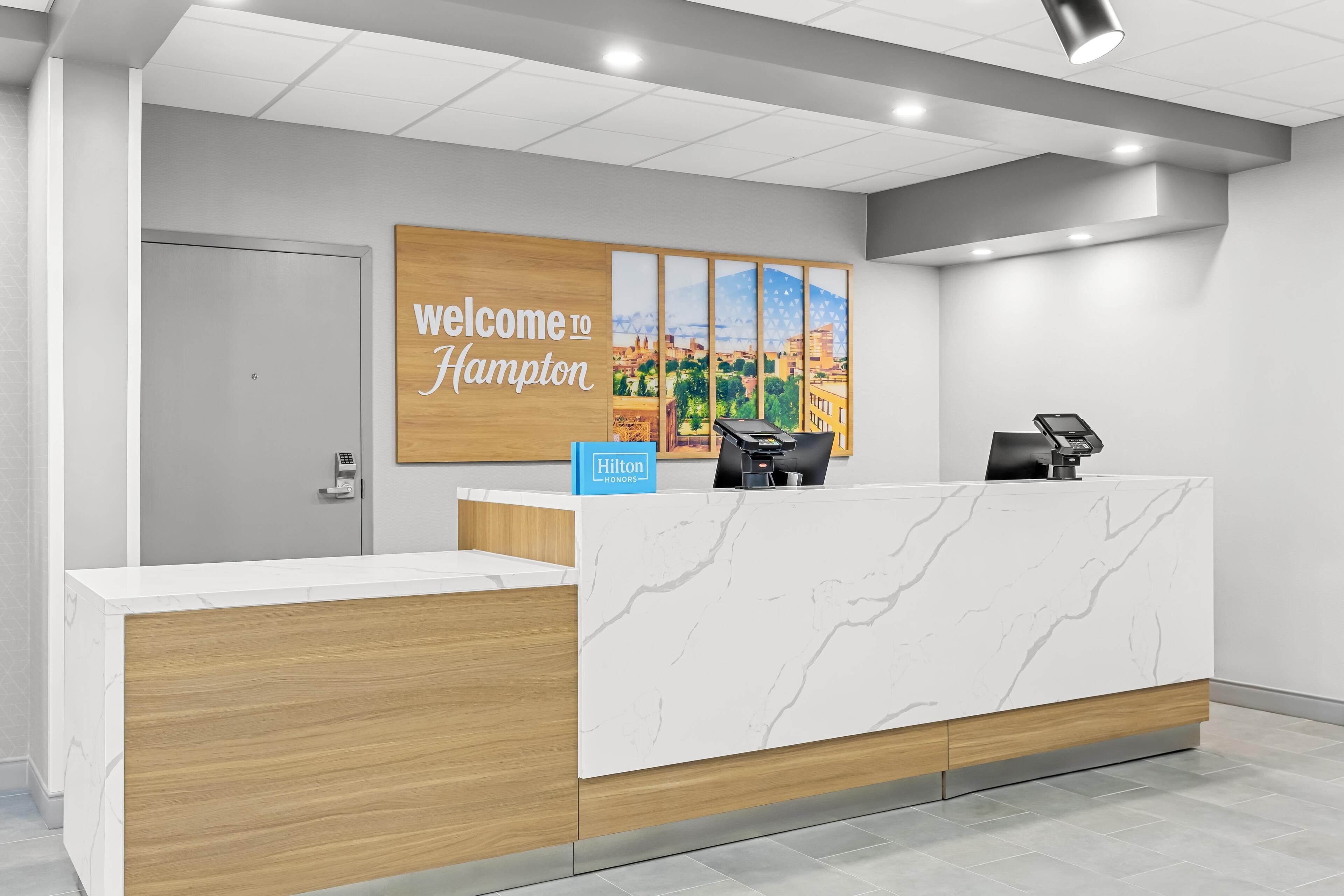 Una moderna recepción de hotel con un mostrador de mármol, un panel de madera con el letrero "Welcome to Hampton" y vistas a la ciudad.