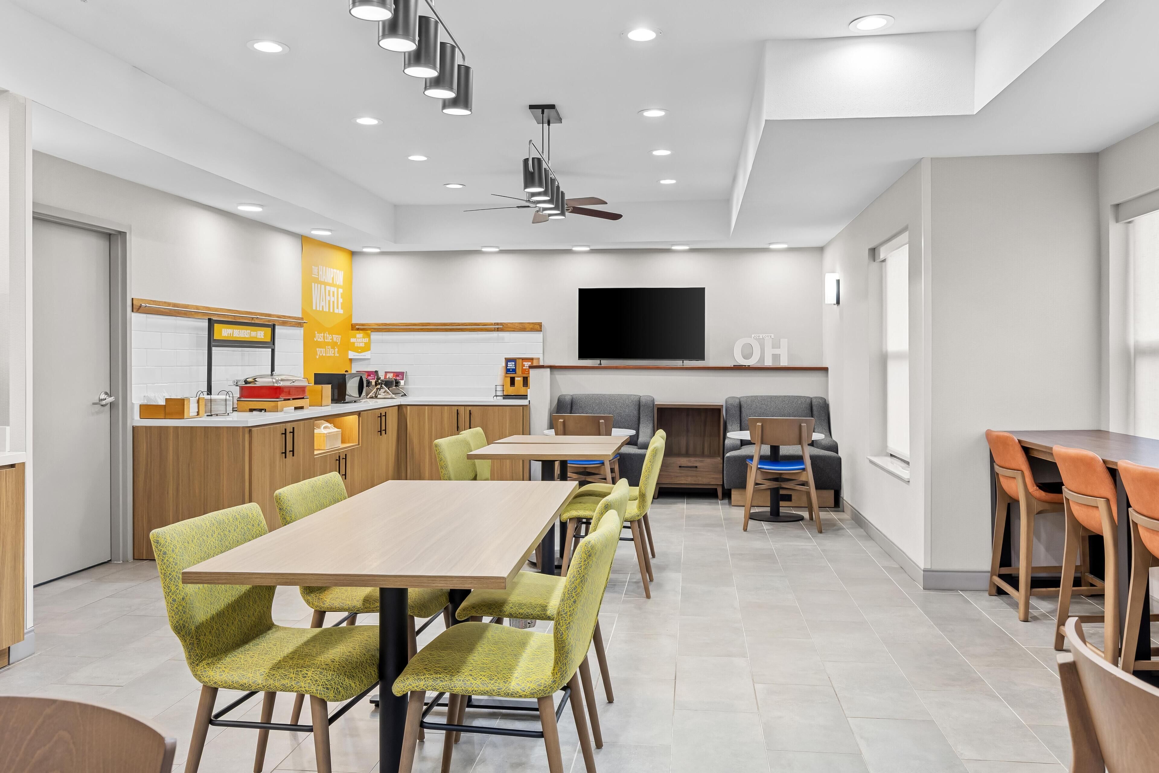 A modern dining area with multiple tables and chairs, a kitchen counter, and a flat-screen TV mounted on the wall.