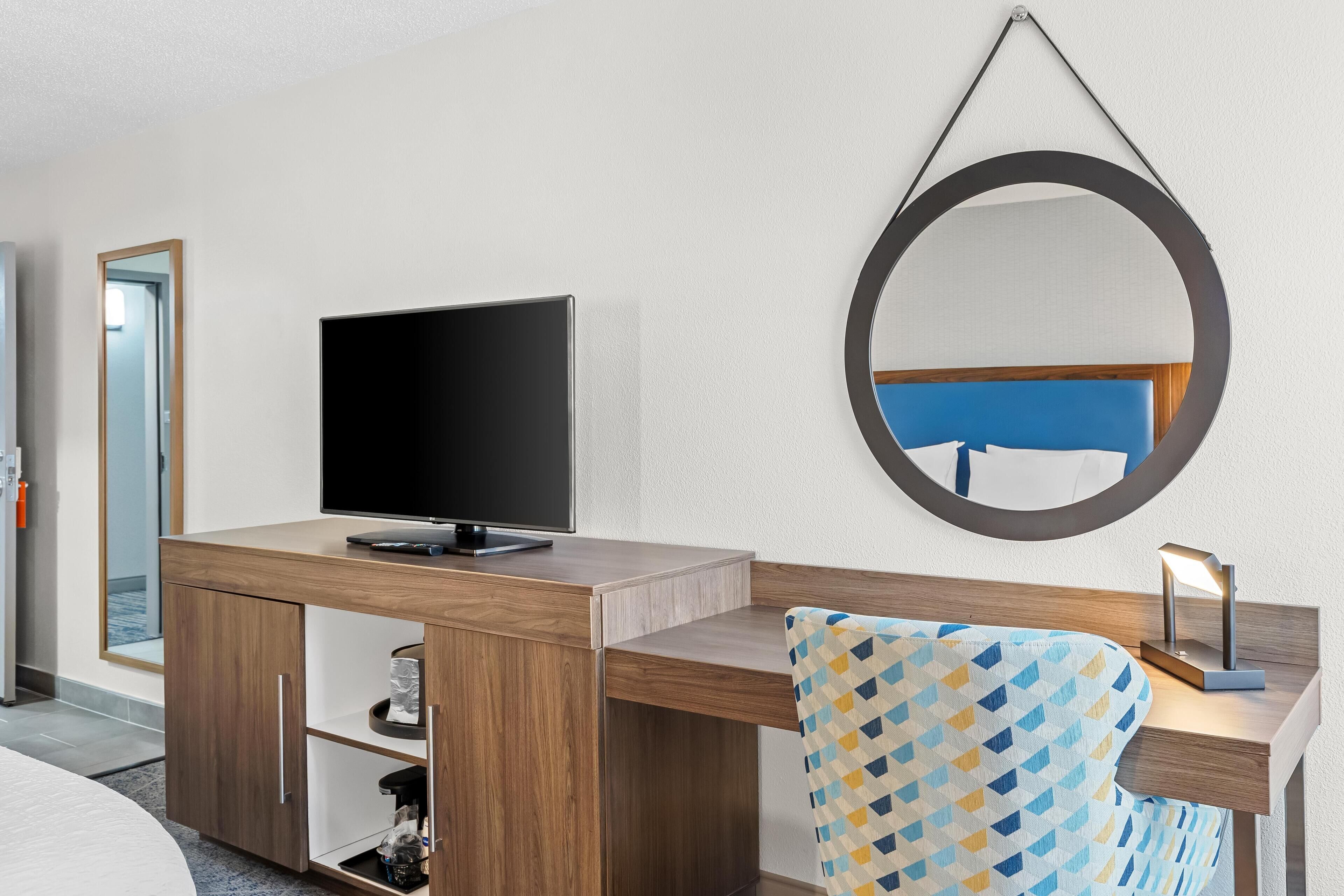 Une chambre d’hôtel avec un bureau en bois, un téléviseur à écran plat, un miroir et un fauteuil à motifs.