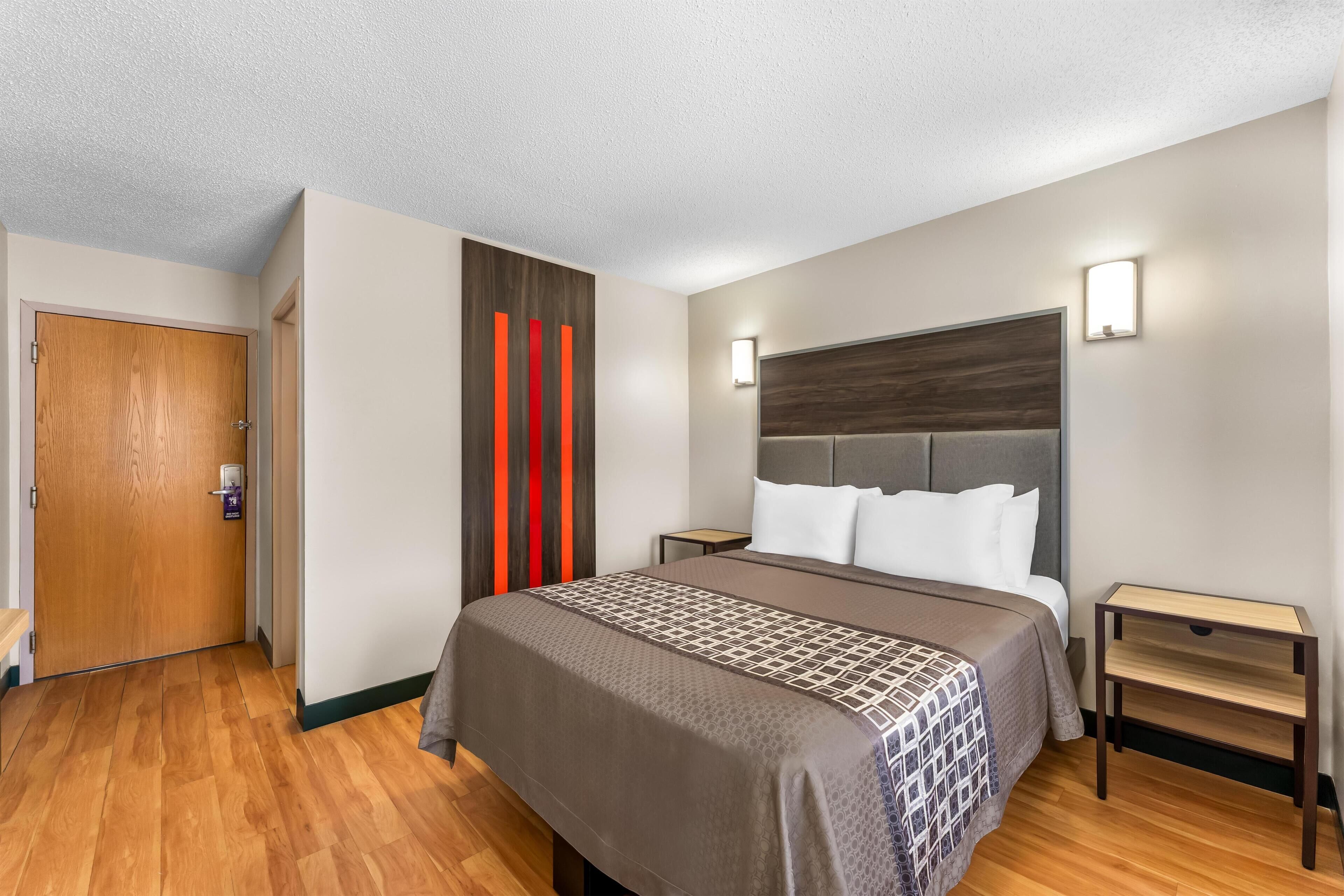 A hotel room with a wooden floor, a bed with a patterned bedspread, a headboard, bedside tables, and a door with a lock.