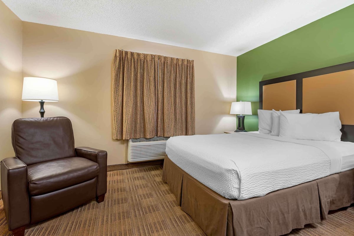 A hotel room with a bed, two bedside lamps, a brown leather chair, a large window with curtains, and a green accent wall.