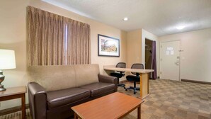 Habitación de hotel con un sofá de cuero marrón, una mesa de centro de madera, un escritorio con dos sillas, una cortina estampada y un cuadro en la pared.