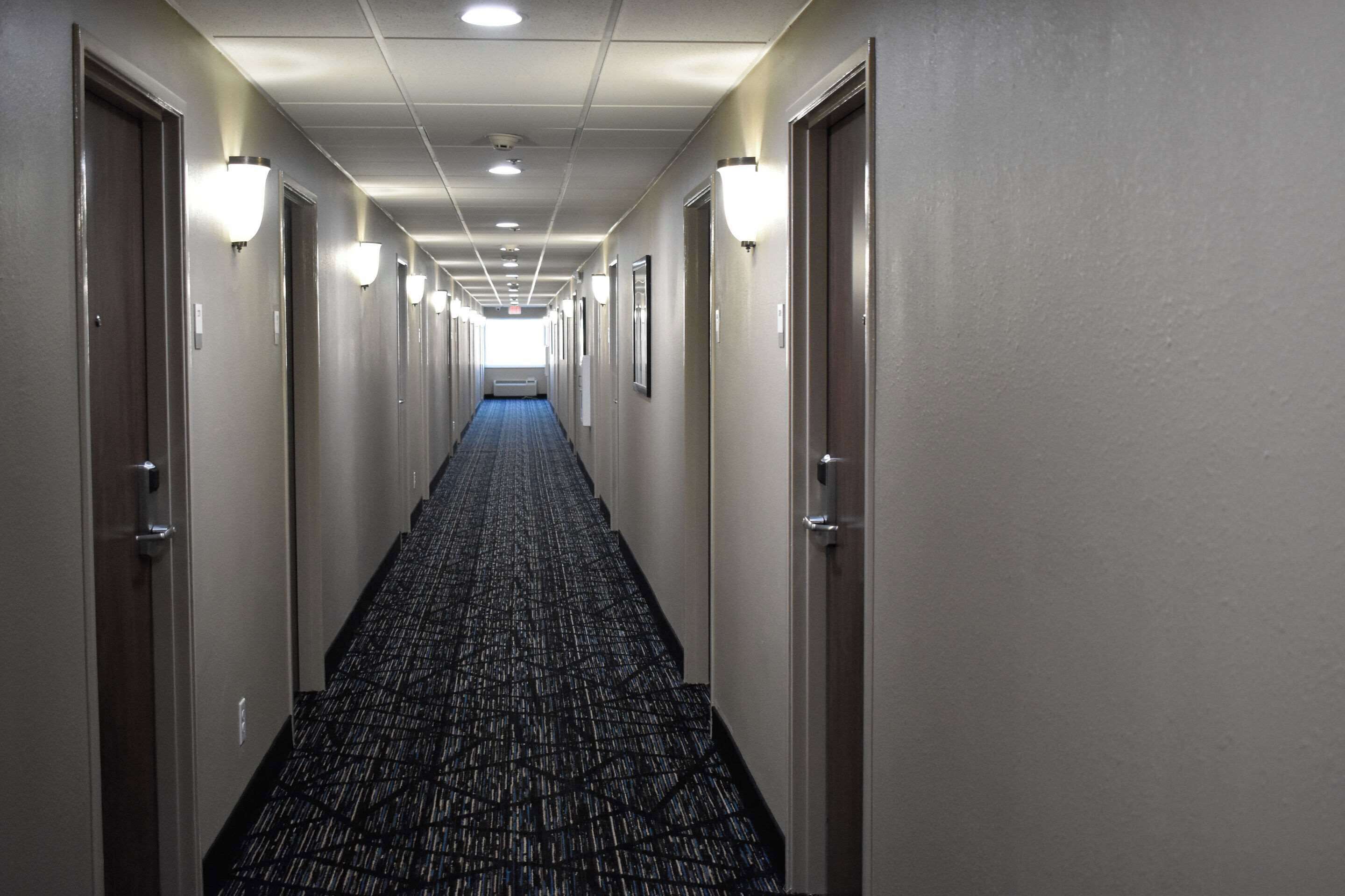 Un pasillo largo y estrecho en un hotel, con suelos alfombrados, puertas a ambos lados y luminarias fijadas a la pared.
